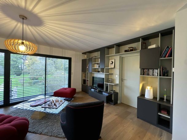 Salon contemporain avec bibliothèque sur mesure encadrant une porte, mobilier noir et gris avec étagères ouvertes, lumière naturelle grâce à de grandes baies vitrées et décoration moderne, incluant une table basse et des fauteuils design.