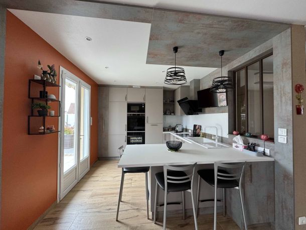 Cuisine moderne avec mur orange accentué, îlot central blanc avec tabourets design, sol en bois clair et luminaires suspendus au-dessus du plan de travail, combinant style contemporain et convivialité.