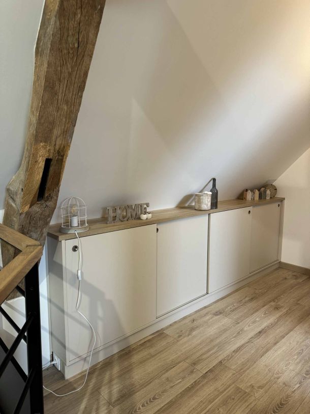 Meuble bas sur mesure en finition blanche et bois clair, installé sous un toit mansardé, avec des éléments de décoration et une poutre apparente en bois rustique.